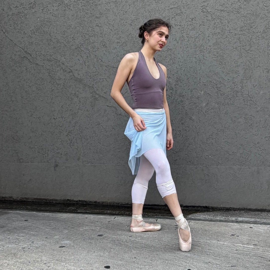 A woman in ballet attire, including a purple tank top, white leggings, a light blue skirt, and pointe shoes, posing against a textured gray concrete wall.