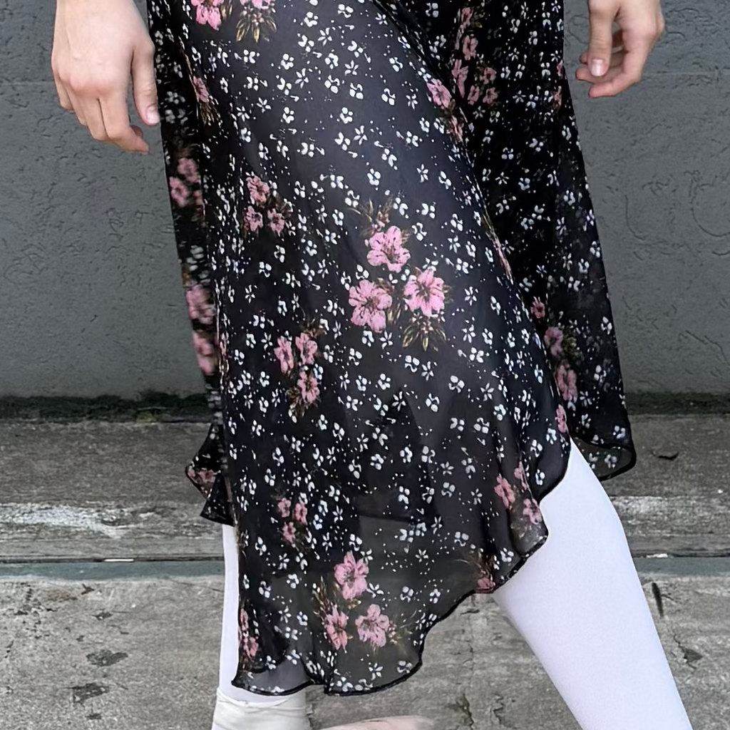 Woman in a purple top and floral skirt standing against a gray wall.