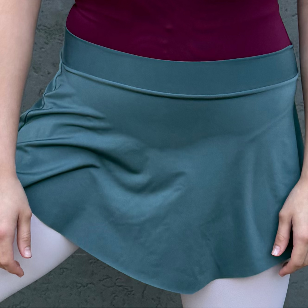 Person wearing a purple top and teal skirt with white tights and ballet shoes against a gray wall.