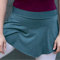 Person wearing a purple top and teal skirt with white tights and ballet shoes against a gray wall.