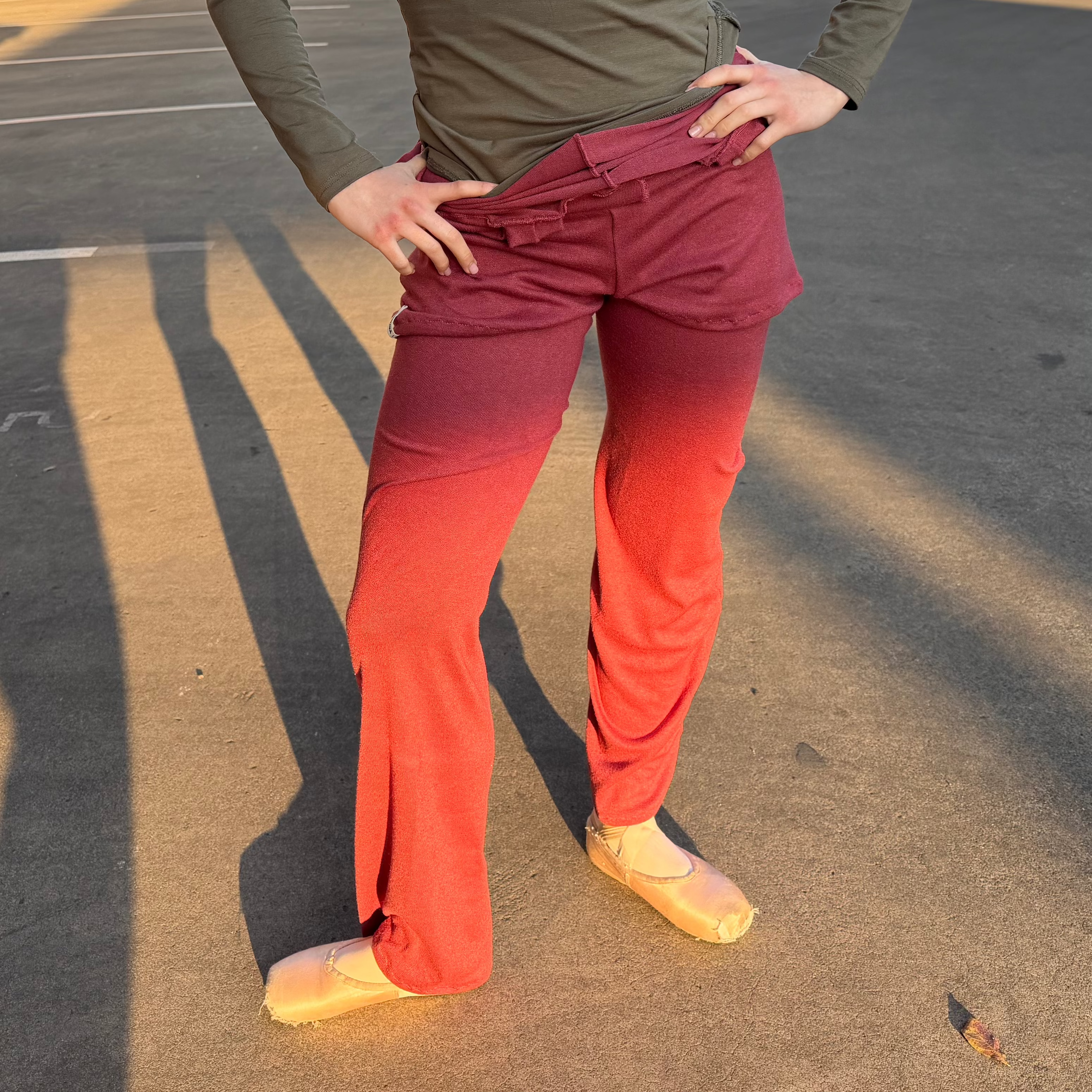A woman stands outdoors in a parking lot wearing a fitted olive green long-sleeve top and high-waisted, wide-leg dance pants with a red-to-orange gradient. She is wearing light tan ballet slippers.