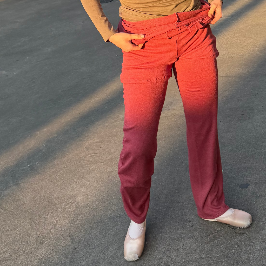 A woman standing in an outdoor concrete parking structure wearing a fitted olive-brown long-sleeve top, bright pinkish-red pants, and white sneakers.