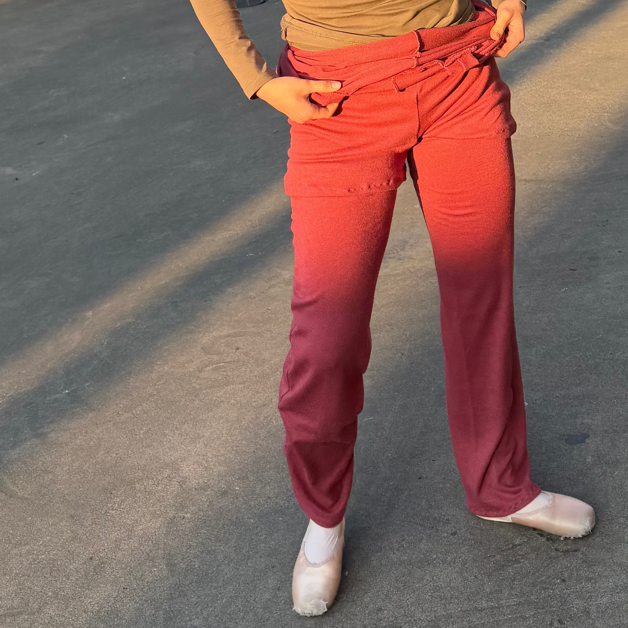 A woman standing in an outdoor concrete parking structure wearing a fitted olive-brown long-sleeve top, bright pinkish-red pants, and white sneakers.