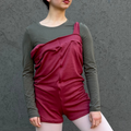 A young ballerina posing in a studio setting against a textured gray wall, wearing a burgundy ruffled sleeveless jumpsuit over a dark gray long-sleeve top, white tights, and pointe shoes.