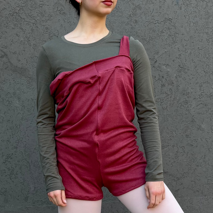 A young ballerina posing in a studio setting against a textured gray wall, wearing a burgundy ruffled sleeveless jumpsuit over a dark gray long-sleeve top, white tights, and pointe shoes.