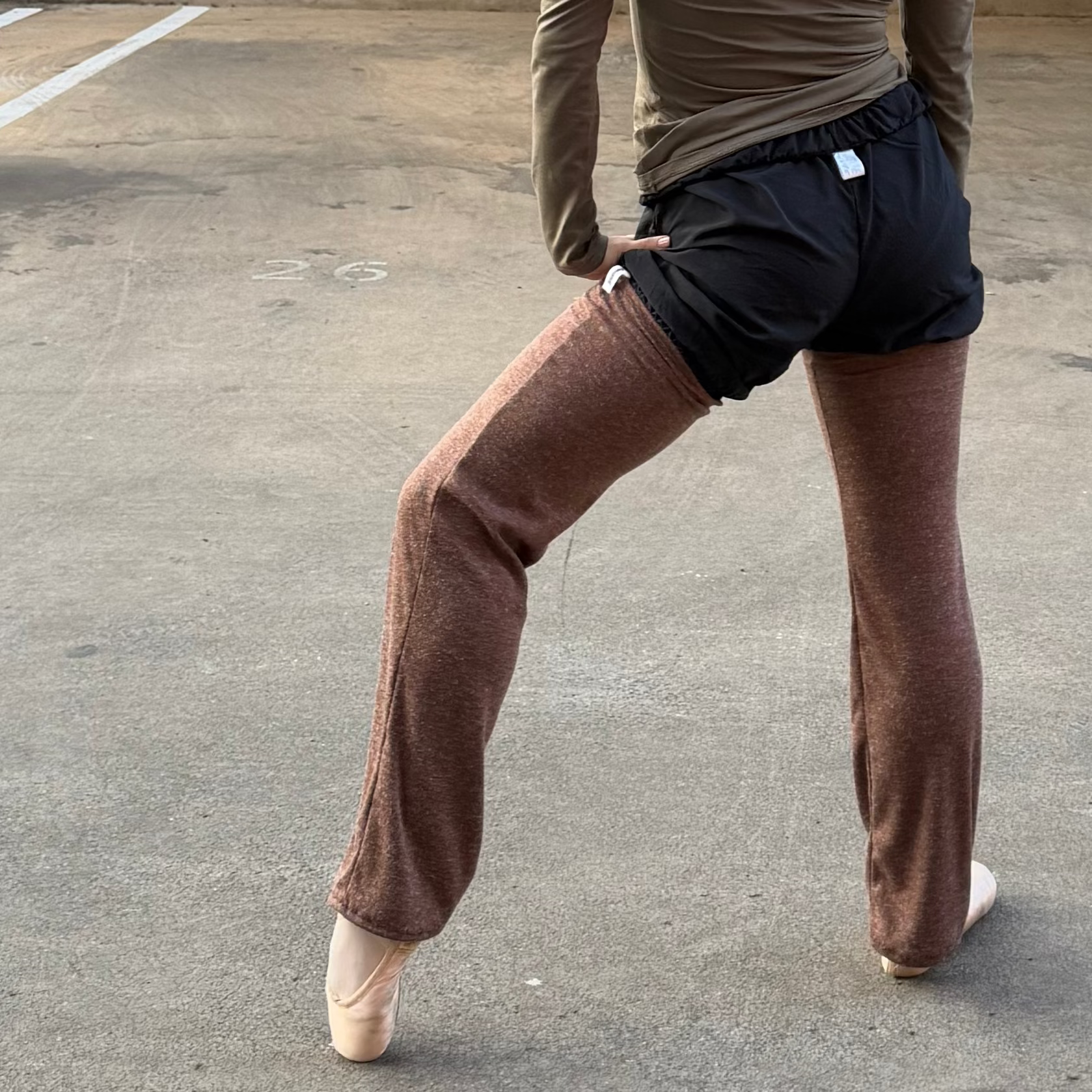 A woman in a dance pose wearing a muted olive green long-sleeve top, black shorts, and brown leggings in an outdoor concrete parking structure.