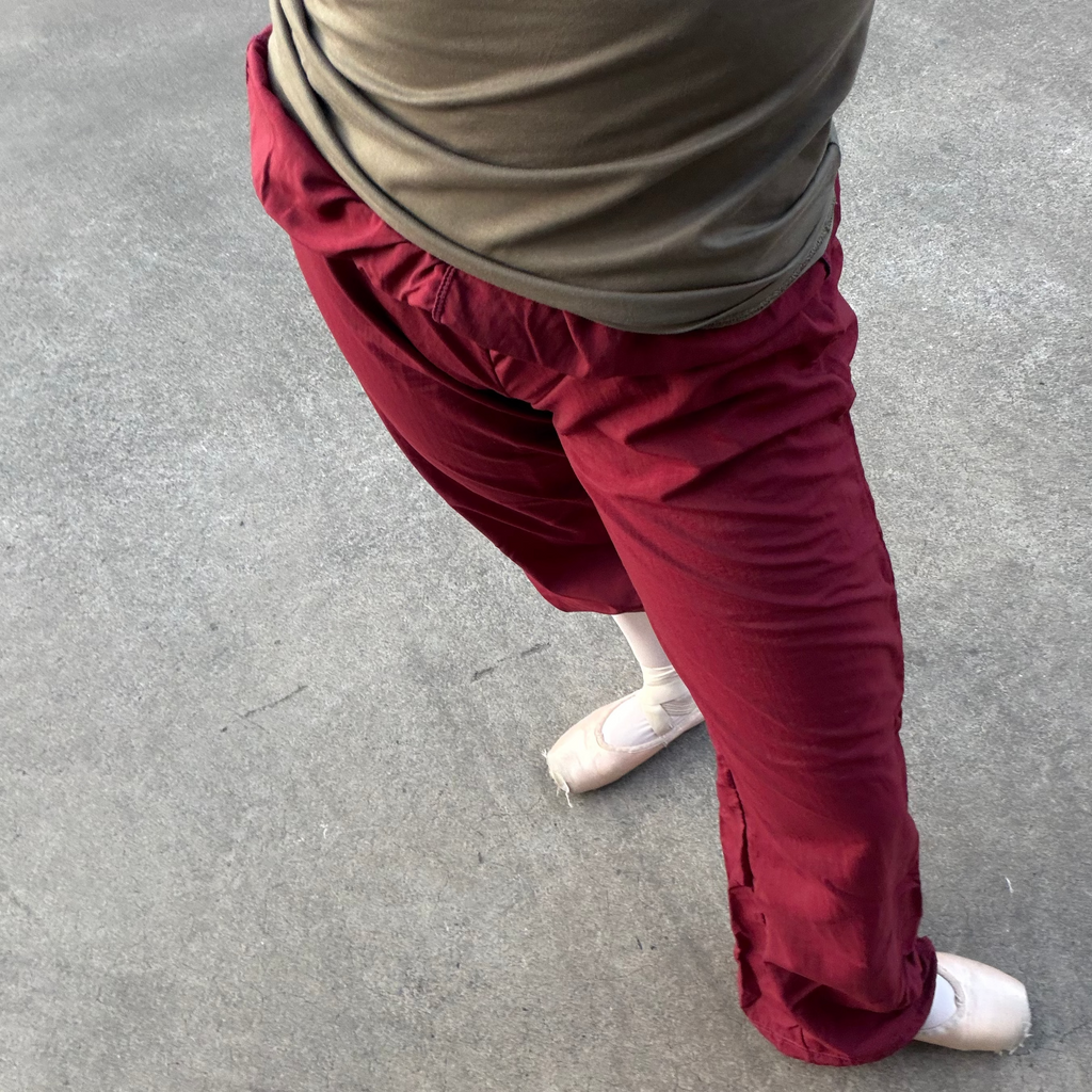 A person wearing loose burgundy-red pants and a taupe-beige top, standing barefoot on a gray concrete surface.