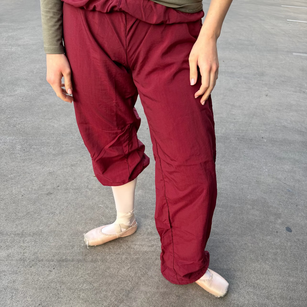 A person wearing loose burgundy dance tights or cropped pants with a gathered elastic waist, paired with a muted green top and pink pointe shoes, standing on a gray paved surface.